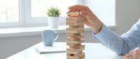 Man in light blue shirt carefully placing wooden block on stacking tower game at office desk, representing business risk, strategy, and decision-making concepts for corporate training.