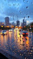 Raindrops on window glass with blurred city skyline and street lights creating bokeh effect at dusk, moody urban scene through wet surface.