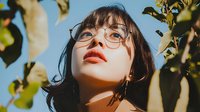 Young Asian woman with round glasses and bright orange lipstick gazing upward among green leaves against blue sky, creating a dreamy outdoor portrait.