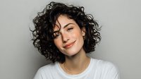 Young woman with curly dark hair and natural makeup smiling confidently against gray background, wearing casual white t-shirt.