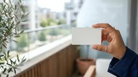 Man with dark skin tone holding blank white business card near window with city view and potted plant, suitable for corporate branding and professional networking concepts.