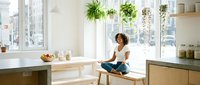 Young woman with curly dark hair, sitting on wooden stool by bright kitchen window with hanging plants, wearing casual white top and dark pants, relaxed home interior setting.