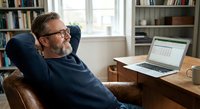 Relaxed mature Caucasian businessman with beard and glasses leaning back in leather chair at home office desk with laptop displaying spreadsheet data.