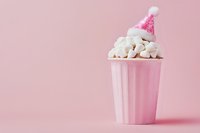 Pink striped paper cup filled with white marshmallows topped with a small pink party hat on solid pink background. Use for celebration, dessert, candy, and festive event marketing materials.