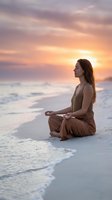 Young Caucasian woman meditating in lotus pose on sandy beach at sunset, waves gently touching shore, warm golden sky, mindfulness concept.