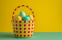 Yellow and red polka dot basket filled with colorful Easter eggs on green surface against vibrant yellow background, minimal holiday composition.