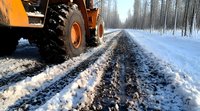 Heavy machinery wheel on snowy forest road during winter maintenance, close-up view of tire tracks in fresh snow with pine trees lining rural path.