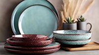Rustic ceramic dinnerware set featuring turquoise and terracotta plates and bowls with speckled texture, arranged on wooden surface with dried pampas grass decor.