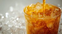 Golden liquid pouring into glass filled with ice cubes creating refreshing cold drink with bubbles and splash, close up view on blurred background.