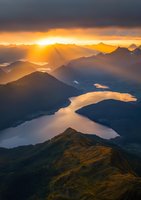 Dramatic sunset over mountain landscape with golden light rays breaking through clouds, illuminating winding lake and peaks in warm orange glow. Aerial wilderness view.