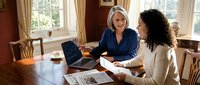 Two professional businesswomen collaborating at wooden table in bright office space, reviewing documents and using tablet for meeting or consultation.