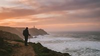 Hiker with backpack standing on coastal cliff at sunset overlooking ocean waves and lighthouse on distant headland, adventure travel and outdoor exploration.
