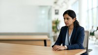 Young South Asian woman in professional navy blazer seated at wooden desk in modern office environment, suitable for corporate, business, and workplace concepts.