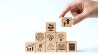 Hand placing wooden cube with house icon completing pyramid of blocks featuring growth symbols for business development concepts.