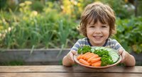 Happy young boy with curly hair smiling at wooden table with plate of fresh carrots and parsley in garden setting for healthy eating concepts.