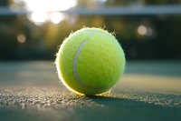 Close-up view of bright yellow tennis ball on court surface at sunset with soft bokeh background, highlighting texture and competitive sports atmosphere.