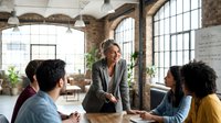 Mature male professional of mixed ethnicity leading business meeting with diverse team in modern loft office space with industrial architecture, large windows, and plants, suitable for corporate training and workplace concepts.