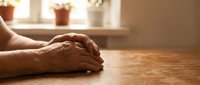 Elderly hands clasped together on wooden table in warm sunlight streaming through window with potted plants creating peaceful home atmosphere.