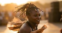 Young African woman with dreadlocks smiling joyfully in warm golden sunset light, expressing happiness and positive emotions in outdoor urban setting.