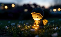 Glowing garden ornament angel figure illuminated at dusk among flowers and grass, creating magical atmosphere with bokeh lights in background.