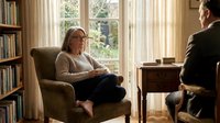 Mature woman with gray hair and glasses sitting in armchair during therapy session with male counselor, home library setting with bookshelves and natural window light.