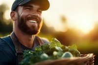 Bearded Caucasian male farmer smiling while holding fresh harvested vegetables in wooden crate during golden sunset hour, organic farming lifestyle concept.