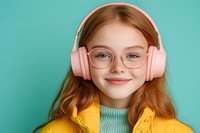 Young redhead girl wearing pink headphones and glasses smiling at camera against turquoise background for music and technology concepts.