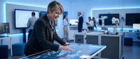 Mature male professional with gray hair and light complexion wearing dark blazer interacts with digital touchscreen interface in modern tech office environment with colleagues and blue lighting.