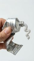 Toothpaste tube being squeezed by hand against white background, showing white paste dispensing from metal container with text label for dental hygiene and oral care products.