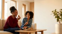 Two African American women laughing together while working on laptop in bright modern living room with natural light from windows and wooden furniture.