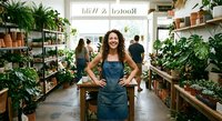 Young woman plant shop owner in denim apron standing confidently among green houseplants in modern nursery store with customers browsing botanical collection.