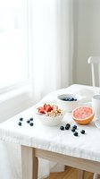 Healthy breakfast table with yogurt granola bowl, fresh strawberries, blueberries, and grapefruit half in bright white morning setting.