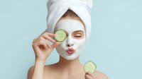 Young Caucasian woman with facial mask holding cucumber slices during skincare routine against light blue background, puckering lips for playful selfie.
