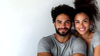 Young diverse couple wearing grey t-shirts smile warmly at camera against white background, showing genuine happiness and connection in casual lifestyle portrait.