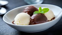 Chocolate and vanilla ice cream scoops served in white ceramic bowl with fresh mint leaf garnish on dark background, close up view of creamy frozen dessert.