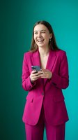 Happy young businesswoman in bright pink suit laughing while using smartphone on turquoise background for corporate communication and social media marketing.
