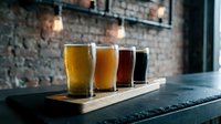 Beer flight tasting set with four glasses displaying different styles from light lager to dark stout on wooden board, craft brewery or pub setting.