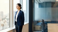 Asian female professional in navy blazer and white shirt standing by office window overlooking cityscape, suitable for corporate business, leadership, and workplace concepts.