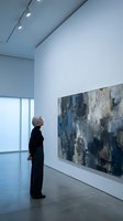 Senior woman contemplating abstract artwork in modern gallery space with track lighting and large windows for cultural appreciation and art viewing.