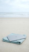 Laptop computer resting on light blue fabric towel on sandy beach with ocean waves in background, representing remote work, digital nomad lifestyle, and vacation productivity.