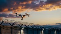 Surveillance drone flying over security fence with barbed wire at dramatic sunset sky for monitoring and protection operations.