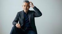 Mature man with gray hair and beard, wearing dark blazer and navy shirt, sitting in confident pose against white background.