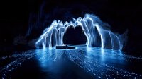 Glowing blue light trails form magical arch over dark water, creating ethereal cave-like structure with reflections rippling across mysterious night landscape.