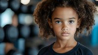 Young African American girl with natural curly hair and expressive eyes gazes directly at camera, wearing black top against blurred bokeh background. Portrait captures innocence and confidence.