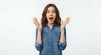 Young Caucasian woman with brown wavy hair showing surprised excited expression with raised hands wearing denim shirt isolated on white background.