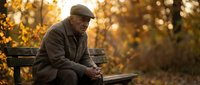Elderly man in warm coat and cap sitting alone on park bench during golden autumn sunset, contemplating in peaceful solitude with bokeh background.