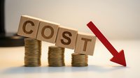 Wooden blocks spelling COST balanced on coin stacks with red arrow pointing down, representing expense reduction and financial savings concept.