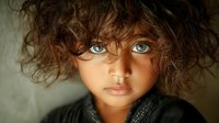 Young child with curly brown hair and striking blue eyes wearing dark clothing against neutral background for emotional storytelling concepts.