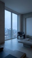 Modern minimalist high-rise apartment interior with floor-to-ceiling windows overlooking urban cityscape, featuring concrete walls, black sculptural armchair, and low platform table for contemporary office or residential design projects.