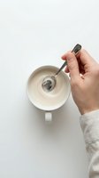 Person holding spoon over white mug with creamy substance, overhead view on neutral background. Suitable for food, cooking, and lifestyle design projects.